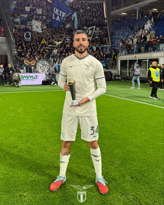 A soccer player in white SS Lazio jersey numbered 34 stands on green field holding silver trophy with both hands, wearing matching shorts and socks, red cleats, stadium seating and crowd in background with blue banners displaying team logo.