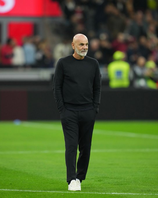 Stefano Pioli stands on a green football field near the sideline with hands in pockets wearing a black long-sleeve sweater black pants and white sneakers surrounded by a blurred stadium crowd and advertising boards in the background