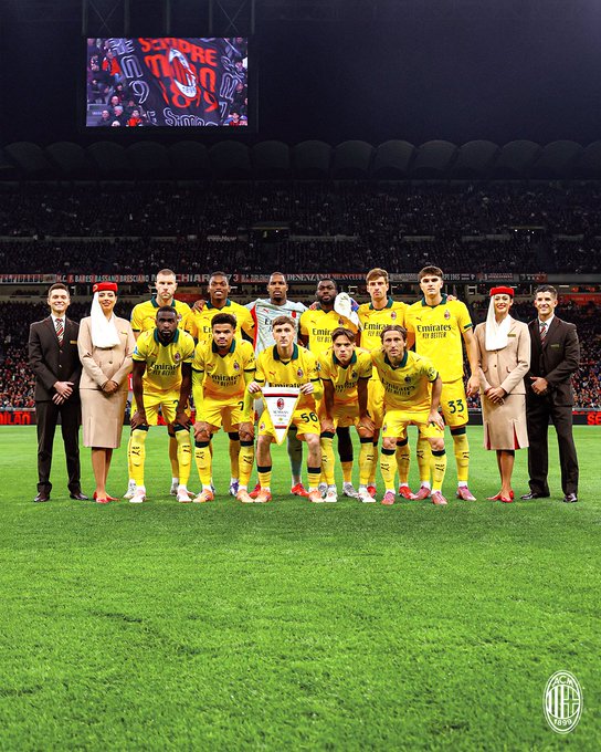 Players in yellow jerseys with black accents and shorts stand in formation on a vibrant green soccer field under stadium lights with a large digital scoreboard displaying red and black Emirates branding in the background coaches in dark suits and light coats flank the group some team members wear red headscarves others hold scarves the AC Milan logo appears at the bottom right