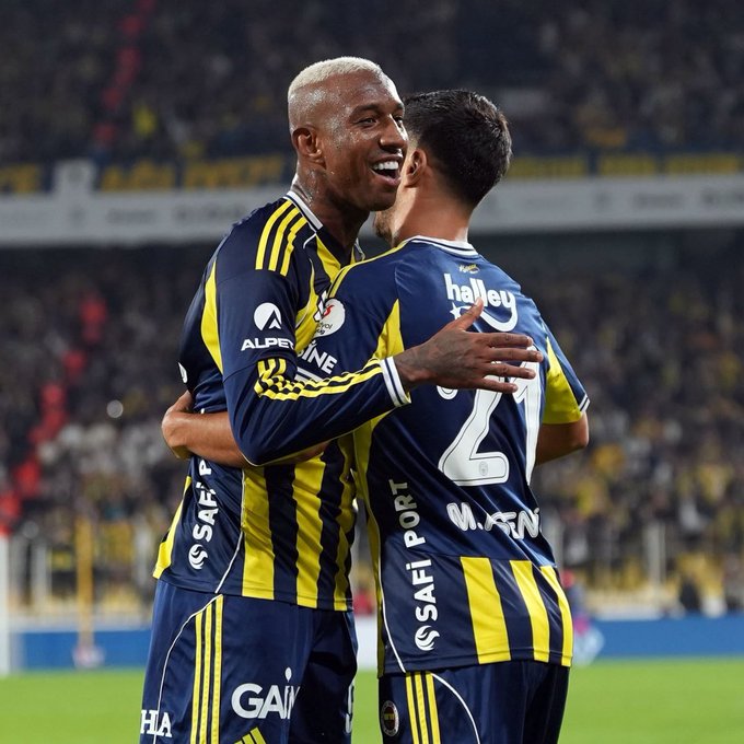 Two male soccer players in yellow and navy Fenerbahce home jerseys with sponsor logos like Safi and Alpine embrace and smile while facing each other on a green stadium field with blurred yellow-seated stands and advertising boards in background one player has short white hair and number 21 on back other has dark hair and number 2 on back