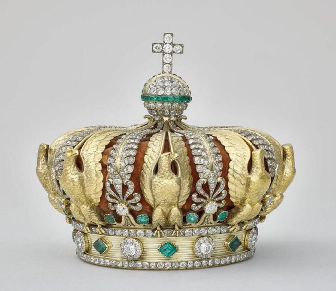 Ornate golden crown with a cross on top featuring diamonds and emeralds, surrounded by detailed filigree work, feathers, and gemstone inlays in a historical style.