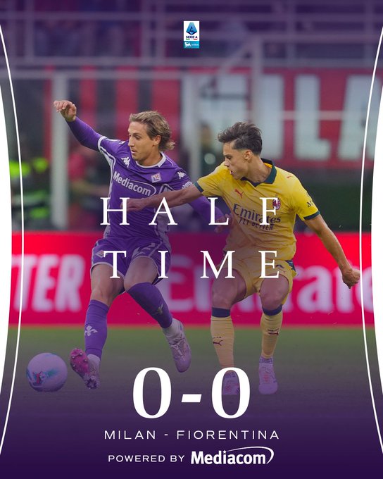 Two soccer players in action on a green field during a match one with long blond hair wearing a purple ACF Fiorentina jersey with white accents and sponsor logos running with the ball while the other with dark curly hair in a yellow AC Milan kit with red and black details pursues the ball stadium seating and advertising boards visible in the background large white text overlay reads HALF TIME in bold letters below it 0-0 score and MILAN FIORENTINA match title at the bottom powered by Mediacom logo in purple