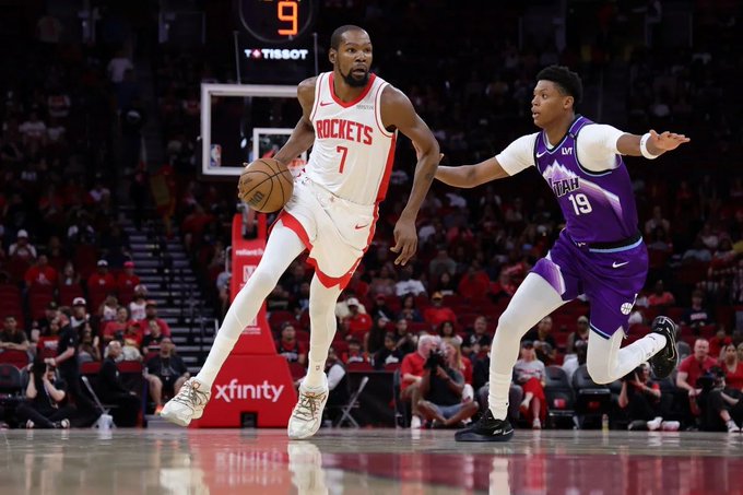 Basketball player Kevin Durant in red Houston Rockets jersey number 7 dribbles the ball while defended by a purple Utah Jazz player number 19 extending an arm, on a court with scoreboard showing time remaining and crowd in stands.