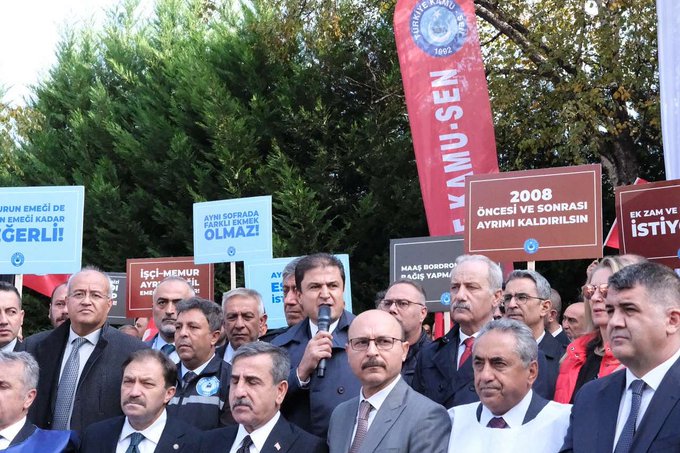 Group of middle-aged men in suits and ties standing outdoors near green trees and flags, one man in glasses holding a microphone and speaking, banners in Turkish reading Egitim-Sen, Ocak 2008, and other text about organization and protests, red and blue flags visible, formal rally setting with participants looking attentive.