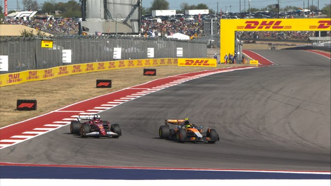 Two Formula 1 race cars navigate a turn on the Circuit of the Americas track during the United States Grand Prix with a red Ferrari car driven by Charles Leclerc leading slightly ahead of a yellow McLaren car driven by Lando Norris on the asphalt surface lined by yellow and white curbs red and white barriers and sponsor banners including DHL in the background under clear skies with spectator stands and fencing visible.