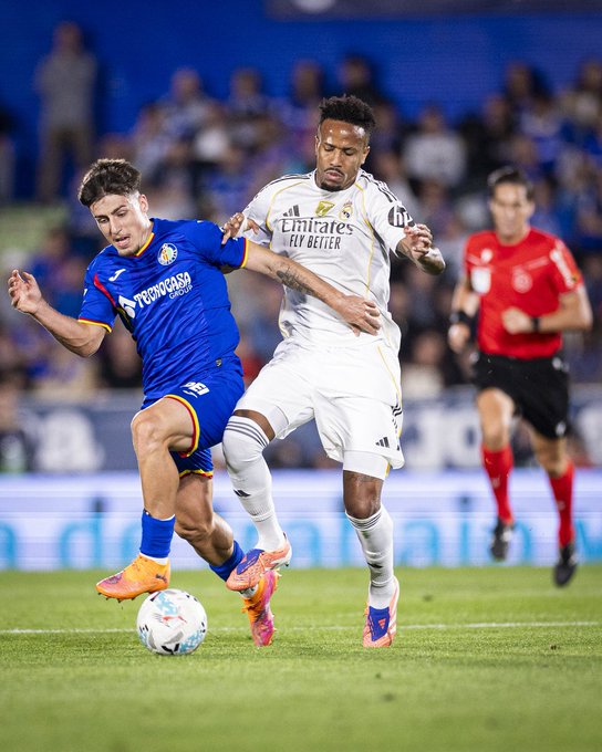 Two soccer players in action on green field during match one in blue Getafe CF jersey with orange boots holding white ball other in white Real Madrid jersey with pink boots referee in red nearby crowd in blue stands background stadium setting