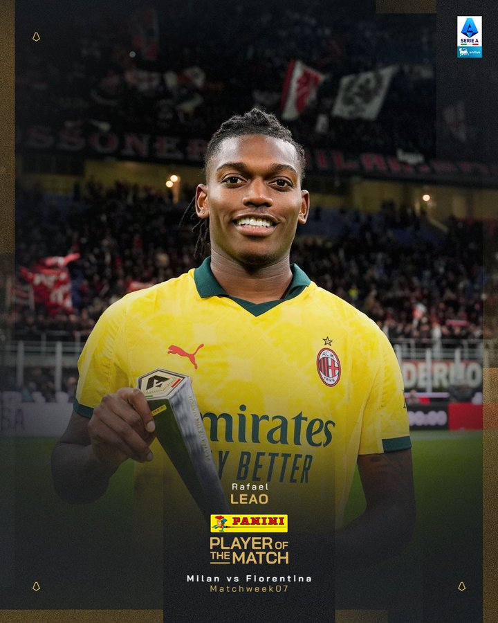 A young Black male soccer player with braided hair smiles while holding a trophy labeled Panini Player of the Match for Milan vs Fiorentina Matchday 07, wearing a yellow AC Milan jersey with Emirates Fly Better sponsor logo, standing on a field with stadium seating blurred in background featuring red and white flags and banners.