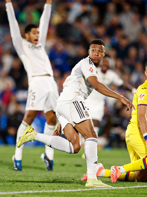 A soccer player in a white Real Madrid jersey with number 17 and black stripes on the sleeves and shorts, wearing green Nike cleats, extends his right arm forward in celebration on a green field during a match, with another teammate in a similar white jersey raising both arms behind him, and opposing players in yellow jerseys visible in the blurred stadium crowd background.