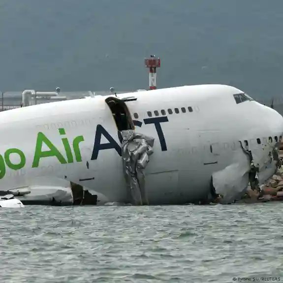 A white Boeing 747 cargo airplane with green Go Air Air lettering on the fuselage lies tilted and partially submerged in shallow water near a runway edge, showing significant damage including a torn-open side section exposing the interior, bent wings, and debris scattered around, with a red control tower visible in the background against a mountainous landscape.