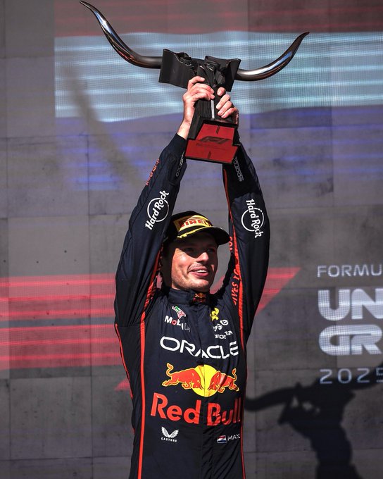 A male driver in a black Red Bull Racing suit with Oracle and Red Bull logos stands on a podium holding a black trophy with curved red horns above, smiling and raising it high. He wears a black cap and the suit has white accents and sponsor patches. The background features a large screen displaying Formula 1 and 202 logos in red and white.