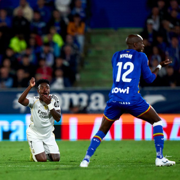Two soccer players on a stadium field during a match one in white Real Madrid jersey number 7 kneeling and raising arms in celebration with open mouth the other in blue Getafe jersey number 12 standing behind with arms raised crowd and blue seating in background.
