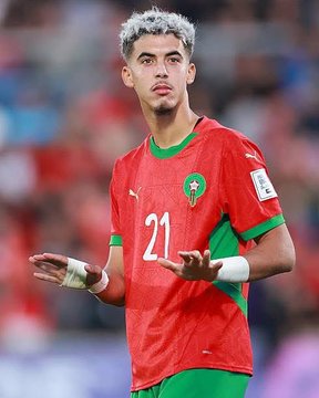 A young male athlete with silver-dyed hair wearing a red and green Morocco national team soccer jersey with number 21 and the team emblem on the chest, standing on a stadium field with blurred crowd in the background, gesturing with both hands extended forward while wearing white wrist wraps.