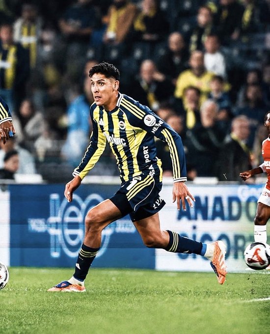Edson Álvarez, a Mexican soccer player with curly dark hair, wears a yellow and navy blue Fenerbahçe jersey with black stripes, navy shorts, and yellow socks while running on a green soccer field during a match. He extends his right arm forward, left leg lifted in motion, with a soccer ball nearby on the grass. Spectators in the stands wear yellow and navy Fenerbahçe scarves and shirts, and advertising boards in Turkish surround the field.
