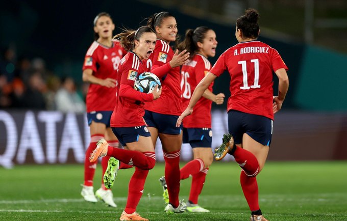 Group of women soccer players from Mexican national team in red Adidas jerseys and shorts running on green grass field during match. Players have numbers like 11 on back with name Rodriguez visible. One player holds FIFA World Cup soccer ball. Background shows stadium seating orange chairs and advertising boards.