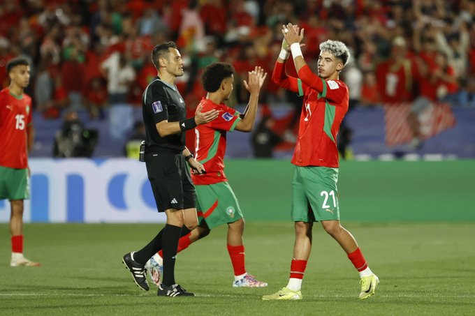 Players in red and green national team jerseys celebrate victory by applauding and raising arms on a green soccer field with stadium seating and crowd in background, referee in black uniform stands nearby, Moroccan flag colors prominent on uniforms and supporters.