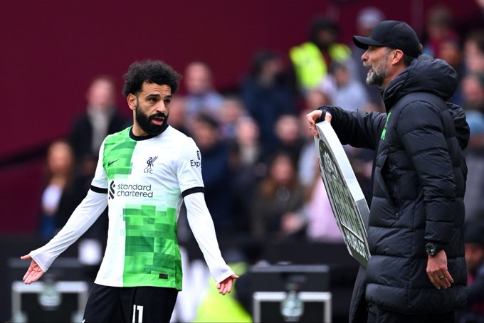 Man with short brown hair and beard wearing light green crewneck sweater sits at wooden table gesturing with open hands while smiling broadly in dimly lit room with lamp and bookshelves behind. Second image shows Mohamed Salah in green and white Fulham kit with number 11 extending arm while Jurgen Klopp in black hooded jacket and cap holds silver trophy plaque on field with blurred crowd and stadium seating in background.