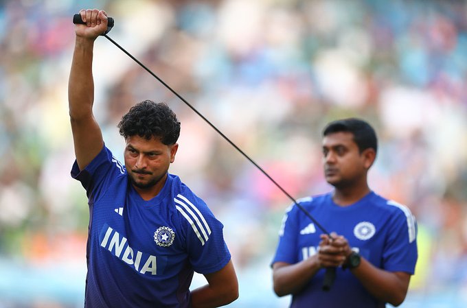 Two male cricket players in blue India national team jerseys with the BCCI emblem stand on a green field during a match, one raising a hand with a resistance band attached to a weight, the other holding a similar band, blurred stadium crowd in the background.