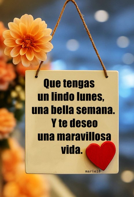 A rectangular wooden sign hangs from a rope against a blue background, adorned with an orange dahlia flower on the left side. The sign displays Spanish text in black letters: Que tengas un lindo lunes, una bella semana, Y te deseo una maravillosa vida. A red heart sticker and signature marie18 appear at the bottom right.