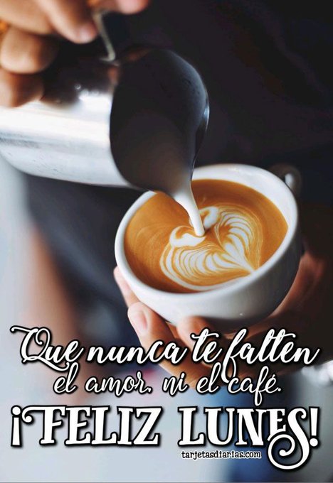 Hands hold a metal pitcher pouring steamed milk into a white ceramic cup containing orange-brown coffee creating a white foam heart shape on the surface. The cup is held by hands against a blurred dark background. Bold white text overlays read Que nunca te fallen el amor ni el café in Spanish followed by Feliz Lunes in larger font.