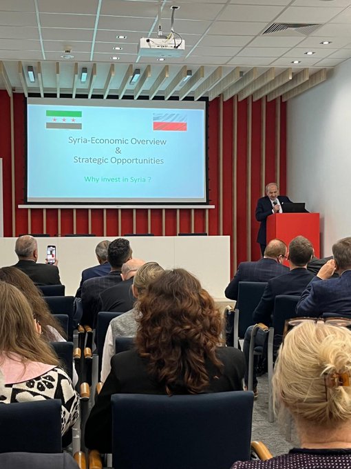 First image shows a group of seven men in business suits standing on a stage with a backdrop displaying Syrian and Polish flags and text Poland-Syria Business Meeting October 2019 one man in the center holds a plaque with a blue ribbon surrounded by others including a man with glasses and another with a beard all positioned formally in front of red curtains and a logo. Second image depicts a conference room with a red backdrop and projector screen showing Syria-Poland Economic Overview and Strategic Opportunities Why Invest in Syria a man in a suit speaks at a red podium while an audience of men and women in business attire seated at tables some with phones listens attentively under ceiling lights and wooden paneling.