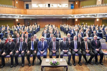 First image shows a man in a suit speaking at a podium on a stage with Turkish flags, the Ministry of National Education emblem, and large screens displaying portraits of Mustafa Kemal Atatürk and Recep Tayyip Erdoğan, banners reading Teftiş Sistemi ve Müfettiş Rollerinin Değerlendirilmesi Çalıştayı, and an audience seated in chairs. Second image depicts an overhead view of a large conference hall with a blue-themed stage, multiple flags including Turkish and organizational banners, rows of attendees in suits seated at tables with water bottles, and ornate wooden paneling on walls. Third image captures a wide group photo of numerous professionals in business attire seated in rows on a stage with a central table holding flowers and water glasses, surrounded by chairs and a backdrop of wooden architecture and lighting.