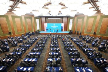 First image shows a man in a suit speaking at a podium on a stage with Turkish flags, the Ministry of National Education emblem, and large screens displaying portraits of Mustafa Kemal Atatürk and Recep Tayyip Erdoğan, banners reading Teftiş Sistemi ve Müfettiş Rollerinin Değerlendirilmesi Çalıştayı, and an audience seated in chairs. Second image depicts an overhead view of a large conference hall with a blue-themed stage, multiple flags including Turkish and organizational banners, rows of attendees in suits seated at tables with water bottles, and ornate wooden paneling on walls. Third image captures a wide group photo of numerous professionals in business attire seated in rows on a stage with a central table holding flowers and water glasses, surrounded by chairs and a backdrop of wooden architecture and lighting.