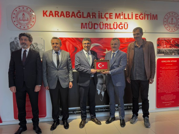 First image displays a formal office setting with a wall sign reading Karabağlar İlçe Milli Eğitim Müdürlüğü, two men in dark suits standing side by side holding a small Turkish flag framed by red borders, one man with glasses and the other with a mustache, behind them Turkish flags and educational plaques including a portrait of Mustafa Kemal Atatürk and text about education and teachers, red and white color scheme dominant. Second image shows five men in formal attire including suits and one in a casual jacket gathered in the same office space under the Karabağlar İlçe Milli Eğitim Müdürlüğü sign, two central figures holding a Turkish flag, others standing nearby with serious expressions, background features Turkish flags, a large Atatürk portrait, and informational panels in Turkish about education initiatives.