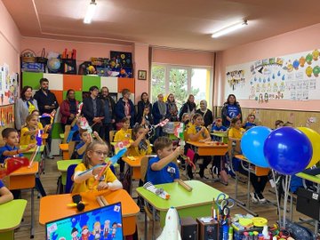 First image shows a large group of adults mostly wearing yellow shirts and some in casual attire standing together in a modern conference room with large windows and a purple backdrop featuring the eTwinning logo, holding a large photo frame of similar group pose, and a large number 2 with eTwinning emblem on the floor in front. Second image depicts a colorful classroom scene with children in orange and yellow shirts sitting at orange desks, holding flags and toys, surrounded by adults in various outfits, with blue balloons, educational posters on walls, and storage cabinets filled with supplies. Third image captures a diverse group of adults in coats and scarves posing together in a school hallway with beige walls, a framed portrait, and a banner in Turkish reading something like Ministry of National Education, near wooden doors. Fourth image presents several women in yellow tops standing on a stage area leading a presentation, gesturing towards a projector screen displaying collaborative learning graphics and eTwinning elements, with tables chairs and laptops in a green-walled meeting room.