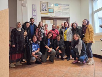 First image shows a large group of adults mostly wearing yellow shirts and some in casual attire standing together in a modern conference room with large windows and a purple backdrop featuring the eTwinning logo, holding a large photo frame of similar group pose, and a large number 2 with eTwinning emblem on the floor in front. Second image depicts a colorful classroom scene with children in orange and yellow shirts sitting at orange desks, holding flags and toys, surrounded by adults in various outfits, with blue balloons, educational posters on walls, and storage cabinets filled with supplies. Third image captures a diverse group of adults in coats and scarves posing together in a school hallway with beige walls, a framed portrait, and a banner in Turkish reading something like Ministry of National Education, near wooden doors. Fourth image presents several women in yellow tops standing on a stage area leading a presentation, gesturing towards a projector screen displaying collaborative learning graphics and eTwinning elements, with tables chairs and laptops in a green-walled meeting room.