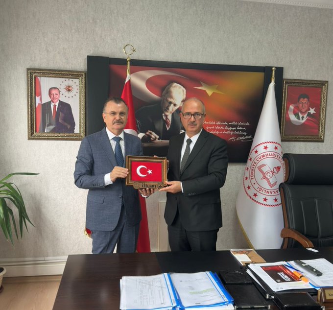 First image displays two men in suits standing behind a desk in an office holding a framed Turkish flag with institutional emblem one man has a mustache and wears a blue suit the other in black suit office features Turkish flags portraits of Mustafa Kemal Atatürk and Recep Tayyip Erdoğan on walls potted plant and documents on desk. Second image shows four people two men and one woman in formal attire with another man behind desk all holding a framed Turkish flag woman wears black top and skirt men in suits office similar with flags portraits potted plant desk items.