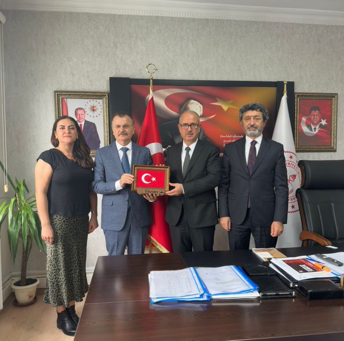 First image displays two men in suits standing behind a desk in an office holding a framed Turkish flag with institutional emblem one man has a mustache and wears a blue suit the other in black suit office features Turkish flags portraits of Mustafa Kemal Atatürk and Recep Tayyip Erdoğan on walls potted plant and documents on desk. Second image shows four people two men and one woman in formal attire with another man behind desk all holding a framed Turkish flag woman wears black top and skirt men in suits office similar with flags portraits potted plant desk items.