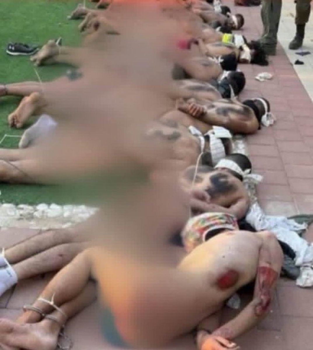This image shows Palestinian detainees stripped of their clothing, blindfolded and bound with zip ties. They appear to have been severely beaten, with visible bruises, cuts and blood on their bodies. They are lying face-down on a paved walkway beside a grassy field, while an