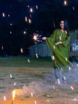 First image: Woman in green embroidered anarkali suit with dupatta sits on ground in park at night, lighting sparkler with smile, surrounded by fireworks and city buildings in background. Second image: Same woman stands holding lit sparkler, green outfit flowing, sparks flying around her joyful expression in grassy area with huts. Third image: Woman holds fountain firework emitting colorful sparks, dressed in green attire, smiling under tree with night sky and distant lights visible.