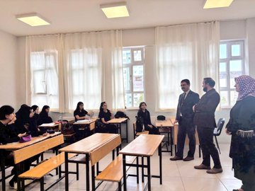 First image shows a group of women in headscarves and professional attire seated around a conference table in a bright classroom with windows and cabinets, engaged in discussion with a man standing and speaking. Second image depicts a man in a suit standing and addressing female students in headscarves seated at wooden desks in a classroom with white curtains and windows. Third image captures a diverse group of male and female students and staff, some in headscarves, standing together in a classroom near desks and a whiteboard. Fourth image features five individuals, including men in suits and women in headscarves, posing in front of a school building entrance with signage for Pen Sosyal Kulup Anadolu Imam Hatip Lisesi.