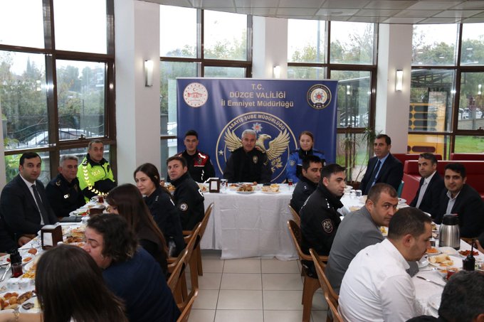 First image shows a large group of people seated at long tables laden with breakfast foods like plates of olives, cheese, bread, and tea in a modern room with large windows and blue banners reading Duzce Vali and Emniyet Mudurlugu. Several attendees wear police uniforms with red armbands and vests, while others are in suits or casual attire including women in headscarves. The setting includes Turkish flags and a central table with the chief presiding. Second image depicts a similar formal breakfast meeting with personnel in blue police uniforms around a table with food items, banners featuring the Duzce Police Department emblem and Turkish eagle, and participants including men and women in professional clothing engaged in discussion.