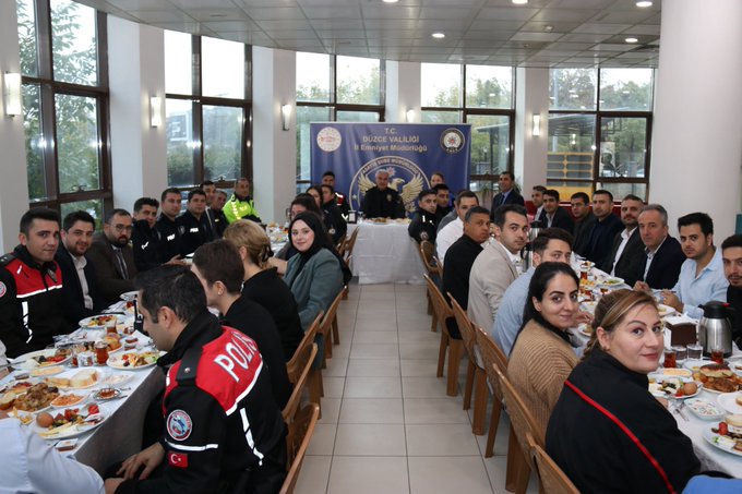 First image shows a large group of people seated at long tables laden with breakfast foods like plates of olives, cheese, bread, and tea in a modern room with large windows and blue banners reading Duzce Vali and Emniyet Mudurlugu. Several attendees wear police uniforms with red armbands and vests, while others are in suits or casual attire including women in headscarves. The setting includes Turkish flags and a central table with the chief presiding. Second image depicts a similar formal breakfast meeting with personnel in blue police uniforms around a table with food items, banners featuring the Duzce Police Department emblem and Turkish eagle, and participants including men and women in professional clothing engaged in discussion.