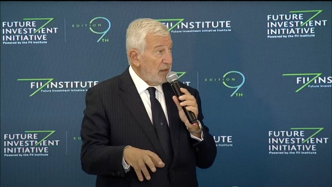 Elderly man with white hair and beard wearing a dark suit and white shirt holds a microphone while speaking on stage. He stands in front of a blue backdrop featuring multiple logos including FII Institute repeated several times and Future Investment Initiative with edition 9 indicators. The setting appears to be a formal press conference event.