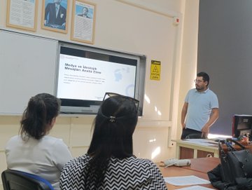First image shows a training room with participants seated at computers around tables listening to a standing facilitator near a large screen displaying educational content. Second image depicts a classroom with two women facing a projector screen showing text about digital media literacy while a male instructor stands nearby. Third image features a group of diverse participants including men and women some wearing headscarves sitting at tables with laptops and engaging in a session with colorful quadrants on a screen. Fourth image captures a male instructor in a yellow classroom pointing at a smartboard displaying a red interface with user profiles while a female participant in a green raincoat stands attentively nearby.