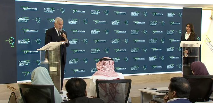 Conference room with blue backdrop displaying Future Investment Initiative logos and PRI logo. Man in dark suit speaks at podium on left wearing microphone. Woman in pink outfit stands at right podium. Audience members in formal attire including some in traditional Saudi garments sit in chairs facing stage. Additional person in suit visible at far right podium.