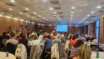 First image shows a large conference room with wooden panel walls and ceiling lights where numerous participants including men and women in casual professional clothing sit at round tables covered with white cloths and bottles of water listening to a speaker on stage with a projector screen displaying content behind them a camera on a tripod records the event doors lead to adjacent rooms. Second image depicts a similar room setup with two projector screens one showing a video call with a woman speaking the other displaying presentation slides participants at tables engage in discussion with notebooks and drinks some wear lanyards. Third image captures a smaller meeting area with a large screen showing a colorful grid chart labeled in Turkish participants including women in headscarves and various attire sit around tables with laptops and documents a woman in blue stands presenting. Fourth image features a group of about 20 diverse participants men and women of various ages and ethnicities standing and sitting in front of a projector screen displaying Code Change World in English and Turkish they pose for a photo some smiling others in professional outfits like shirts and dresses the room has wooden walls and chairs.