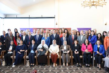 First image shows a large group of formally dressed men and women including some in headscarves standing together in front of purple banners in an indoor hall with wooden floors and hanging lights. Second image depicts a man in a dark suit speaking at a podium with microphones in front of red and purple banners featuring Düzce University and Women Muhtars Support logos. Third image displays rows of seated attendees in business attire including suits and headscarves around tables with a vase of red roses in a room with Turkish flags and large windows. Fourth image captures multiple round tables with groups of women in various outfits including headscarves discussing over notebooks and laptops in a spacious hall with Turkish flags and purple banners.