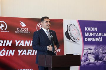 First image shows a large group of formally dressed men and women including some in headscarves standing together in front of purple banners in an indoor hall with wooden floors and hanging lights. Second image depicts a man in a dark suit speaking at a podium with microphones in front of red and purple banners featuring Düzce University and Women Muhtars Support logos. Third image displays rows of seated attendees in business attire including suits and headscarves around tables with a vase of red roses in a room with Turkish flags and large windows. Fourth image captures multiple round tables with groups of women in various outfits including headscarves discussing over notebooks and laptops in a spacious hall with Turkish flags and purple banners.