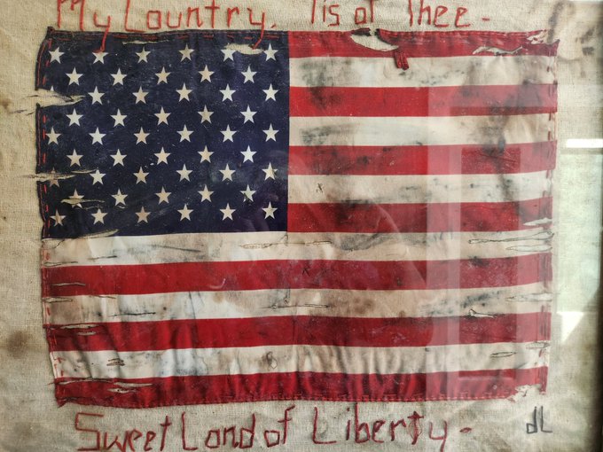 A tattered American flag displayed in a frame, featuring 13 red and white stripes and a blue canton with white stars. Hand-stitched text at the top reads My country tis of thee in faded red letters. Below the flag, embroidered text reads sweet land of liberty in cursive script.