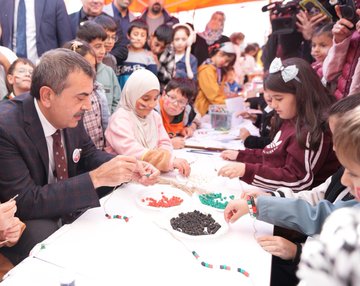 First image shows a man in a suit shaking hands with young girls in school uniforms at a table covered with colorful crafts like bracelets ribbons and beads in a school courtyard with brick buildings and people in headscarves nearby. Second image depicts a large group of children and adults including suited men standing together in front of a school building with Turkish flags some children holding a Palestinian flag and wearing sashes. Third image captures children and a suited man sitting at a white table stringing green red and black beads to make bracelets with craft supplies scattered around in an indoor school setting. Fourth image features a wide group photo of adults in formal attire and children in uniforms posing in front of a modern school entrance with Turkish flags a Christmas tree and a sign reading Özdemir Gürocak İlkokulu.