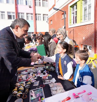 First image shows a man in a suit shaking hands with young girls in school uniforms at a table covered with colorful crafts like bracelets ribbons and beads in a school courtyard with brick buildings and people in headscarves nearby. Second image depicts a large group of children and adults including suited men standing together in front of a school building with Turkish flags some children holding a Palestinian flag and wearing sashes. Third image captures children and a suited man sitting at a white table stringing green red and black beads to make bracelets with craft supplies scattered around in an indoor school setting. Fourth image features a wide group photo of adults in formal attire and children in uniforms posing in front of a modern school entrance with Turkish flags a Christmas tree and a sign reading Özdemir Gürocak İlkokulu.