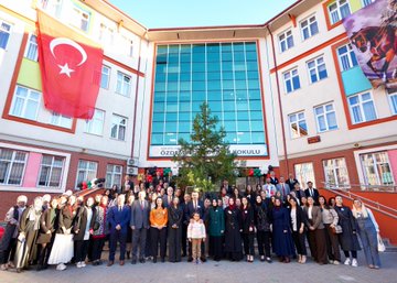 First image shows a man in a suit shaking hands with young girls in school uniforms at a table covered with colorful crafts like bracelets ribbons and beads in a school courtyard with brick buildings and people in headscarves nearby. Second image depicts a large group of children and adults including suited men standing together in front of a school building with Turkish flags some children holding a Palestinian flag and wearing sashes. Third image captures children and a suited man sitting at a white table stringing green red and black beads to make bracelets with craft supplies scattered around in an indoor school setting. Fourth image features a wide group photo of adults in formal attire and children in uniforms posing in front of a modern school entrance with Turkish flags a Christmas tree and a sign reading Özdemir Gürocak İlkokulu.