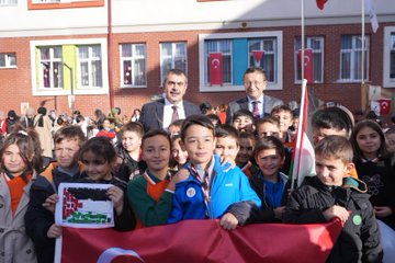 First image shows a man in a suit shaking hands with young girls in school uniforms at a table covered with colorful crafts like bracelets ribbons and beads in a school courtyard with brick buildings and people in headscarves nearby. Second image depicts a large group of children and adults including suited men standing together in front of a school building with Turkish flags some children holding a Palestinian flag and wearing sashes. Third image captures children and a suited man sitting at a white table stringing green red and black beads to make bracelets with craft supplies scattered around in an indoor school setting. Fourth image features a wide group photo of adults in formal attire and children in uniforms posing in front of a modern school entrance with Turkish flags a Christmas tree and a sign reading Özdemir Gürocak İlkokulu.