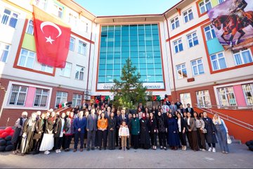 First image shows a group of children and adults gathered under a tent structure with Turkish flags visible some children wearing orange shirts and jackets a man in a suit holding a baby others smiling and posing closely. Second image depicts a man in a suit handing items to children at a table covered with various goods like bottles stickers and crafts in front of a colorful school building with people in headscarves and casual attire standing around. Third image features a large group of children in orange uniforms and adults including a suited man interacting around tables with colorful drawings and boxes near a school entrance area. Fourth image displays a crowd of adults and children in formal and casual clothes standing in front of a school building adorned with Turkish flags a banner reading Özdemir Gürocak İlkokulu and a Christmas tree-like decoration with a ribbon-cutting setup.