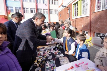 First image shows a group of children and adults gathered under a tent structure with Turkish flags visible some children wearing orange shirts and jackets a man in a suit holding a baby others smiling and posing closely. Second image depicts a man in a suit handing items to children at a table covered with various goods like bottles stickers and crafts in front of a colorful school building with people in headscarves and casual attire standing around. Third image features a large group of children in orange uniforms and adults including a suited man interacting around tables with colorful drawings and boxes near a school entrance area. Fourth image displays a crowd of adults and children in formal and casual clothes standing in front of a school building adorned with Turkish flags a banner reading Özdemir Gürocak İlkokulu and a Christmas tree-like decoration with a ribbon-cutting setup.