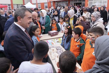 First image shows a group of children and adults gathered under a tent structure with Turkish flags visible some children wearing orange shirts and jackets a man in a suit holding a baby others smiling and posing closely. Second image depicts a man in a suit handing items to children at a table covered with various goods like bottles stickers and crafts in front of a colorful school building with people in headscarves and casual attire standing around. Third image features a large group of children in orange uniforms and adults including a suited man interacting around tables with colorful drawings and boxes near a school entrance area. Fourth image displays a crowd of adults and children in formal and casual clothes standing in front of a school building adorned with Turkish flags a banner reading Özdemir Gürocak İlkokulu and a Christmas tree-like decoration with a ribbon-cutting setup.